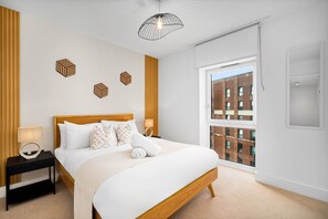 A modern hotel room with a large bed, a nightstand, a lamp, geometric wall art, and a view of a brick building through a window.