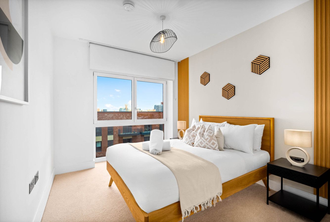 Une chambre d’hôtel moderne avec un grand lit, une table de chevet avec une lampe, une vue sur la ville grâce à de grandes fenêtres et un luminaire au plafond.
