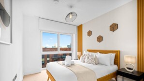 Una habitación de hotel moderna con una cama grande, una mesita de noche con lámpara, vistas de la ciudad a través de amplios ventanales y una lámpara de techo.