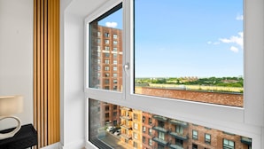 Una habitación con amplios ventanales que ofrecen vistas a un paisaje urbano, una pared de listones de madera y una mesita de noche negra con una lámpara.