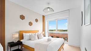 Un dormitorio con una cama grande, mesitas de noche, una ventana con vistas a edificios y un cuadro de pared con motivos geométricos.