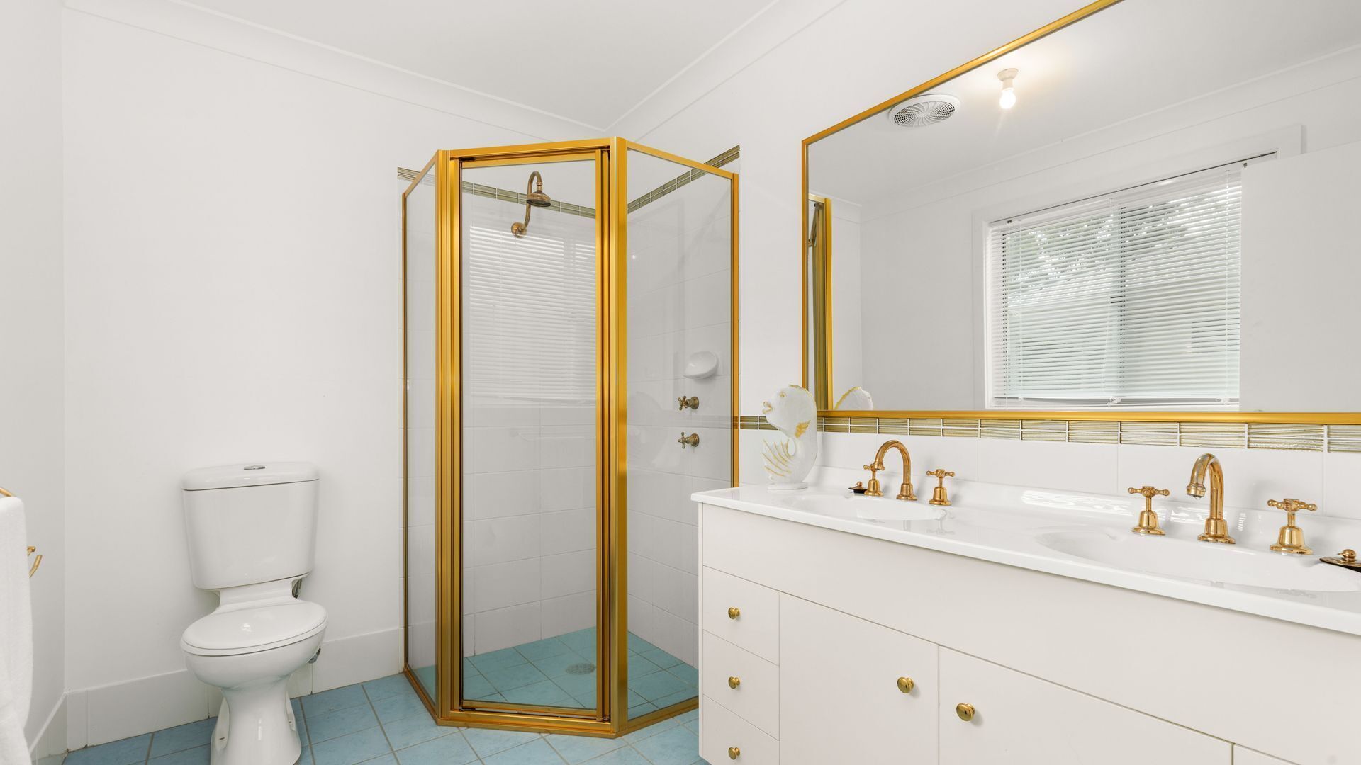 A bathroom with a white toilet, a glass-enclosed shower with gold trim, a white vanity with gold faucets, and a large mirror with a gold frame.