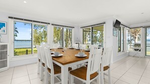 Un coin repas avec une table en bois et des chaises blanches, de grandes fenêtres donnant sur le lac et un téléviseur fixé au mur.