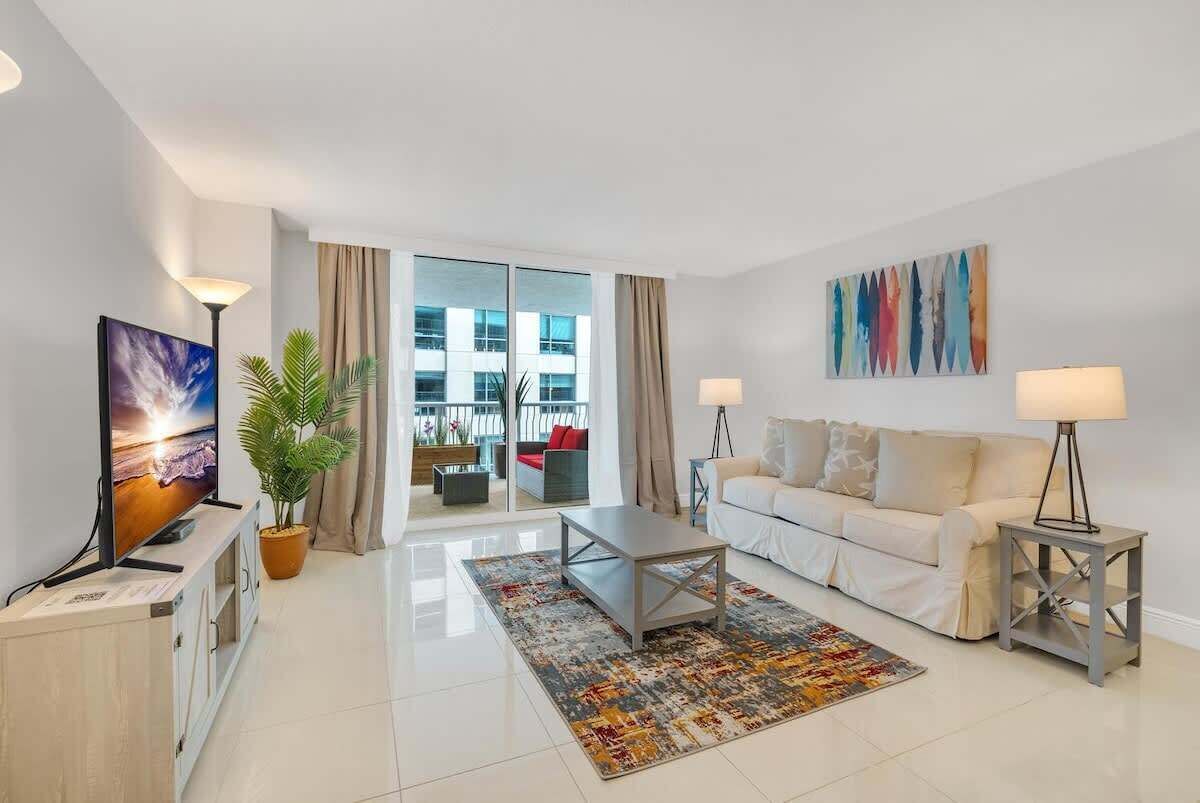 A modern living room with a flat-screen TV, a sofa, a coffee table, and a large window with curtains.