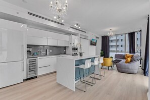 Eine moderne Küche mit weißen Schränken, einer zentralen Kochinsel und Barhocker. Es gibt einen Wohnbereich mit einer Couch und einem Couchtisch.