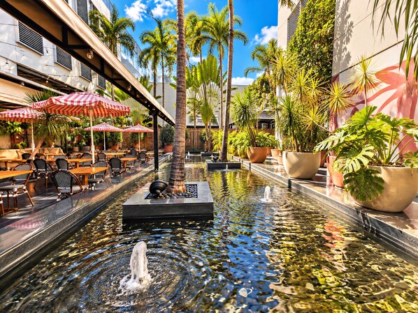 An outdoor dining area with tables and chairs, a water feature with fountains, and potted plants.