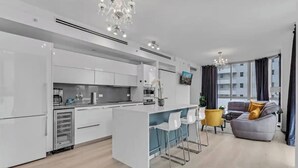 A modern kitchen with white cabinetry, a central island with bar stools, a grey sofa with yellow cushions, and a large window with curtains. - Luxury Miami Beach Apartment with Rooftop Pool (Miami Beach)