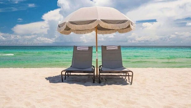 Two beach chairs under a beach umbrella on a sandy beach with clear turquoise water and a partly cloudy sky. - Luxury Miami Beach Apartment with Rooftop Pool (Miami Beach)