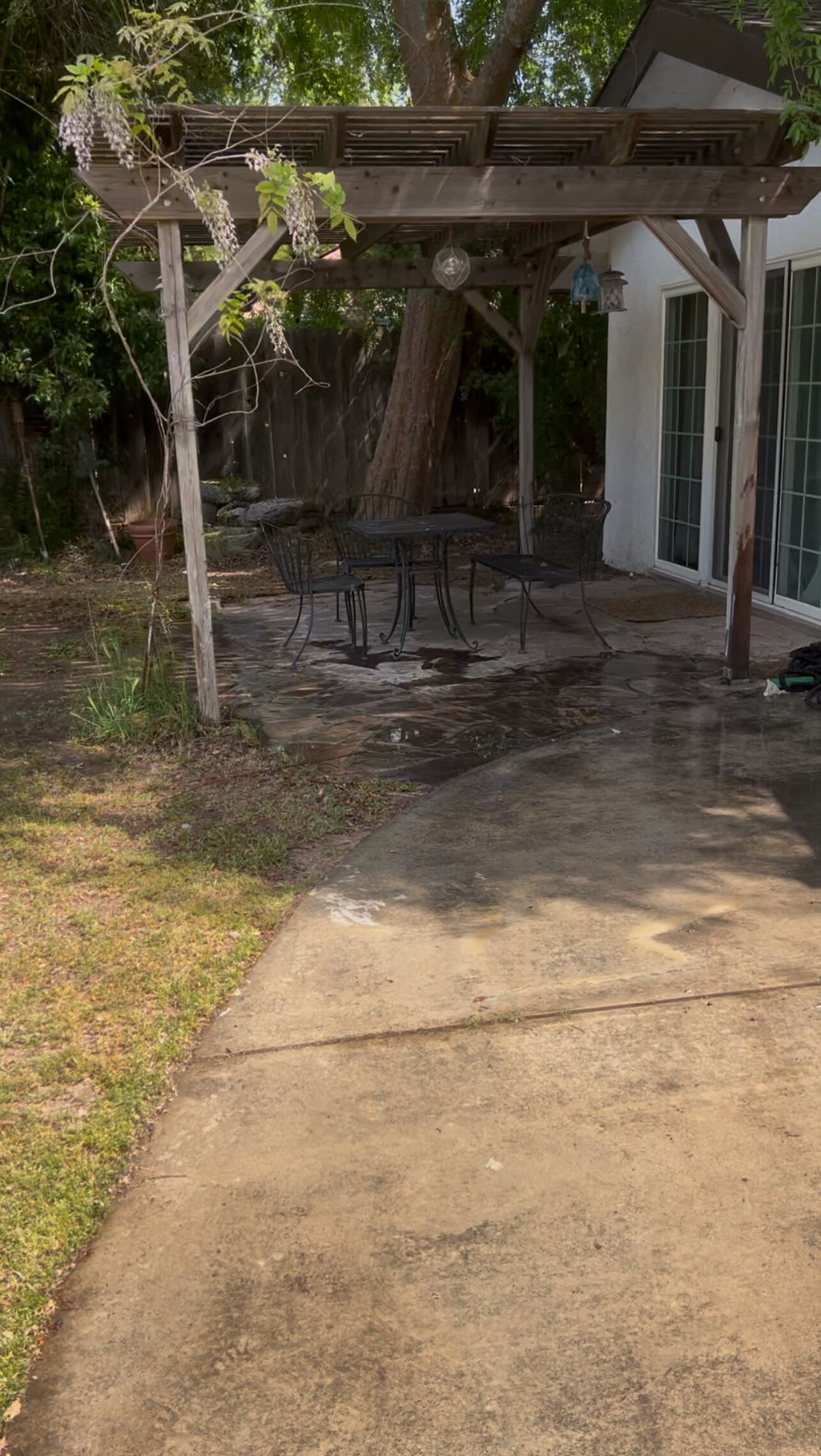 Ein überdachter Patio-Bereich mit Betonboden, umgeben von einer Holzw pergola und einem großen Baum.