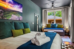 Een hotelkamer met een groot bed, uitzicht op een huis door het raam, en een landschapsschilderij aan de muur.