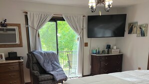 Un dormitorio con cama, ventilador de techo, televisor, cómoda, silla y balcón con cortinas.