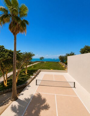 A tennis court with a net, surrounded by palm trees and a view of the ocean.