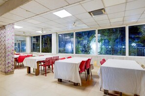 Un comedor con mesas y sillas, ventanas amplias con vistas a árboles, y un techo con baldosas cuadradas y un ventilador.