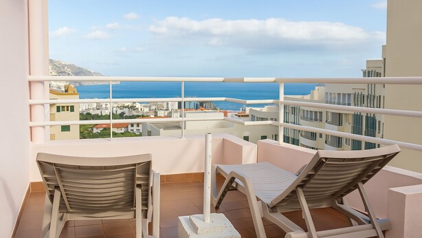 A balcony with two lounge chairs and a table, overlooking a coastal cityscape with buildings and the sea. - Perla do Oceano – Funchal Sea View with XL Terrace (Funchal)
