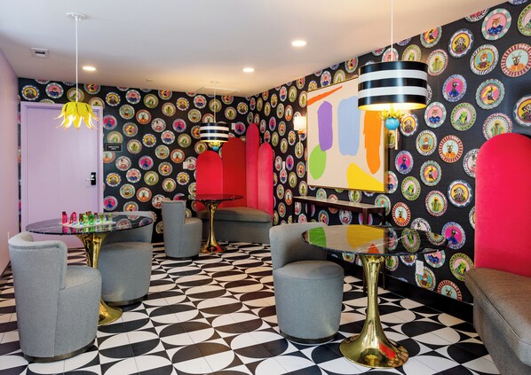 Ein Zimmer mit einem schwarz-weißen geometrischen Bodenmuster, farbenfrohen Wanddekorationen und modernen Möbeln, darunter graue Sessel und ein Glastisch mit goldener Basis.