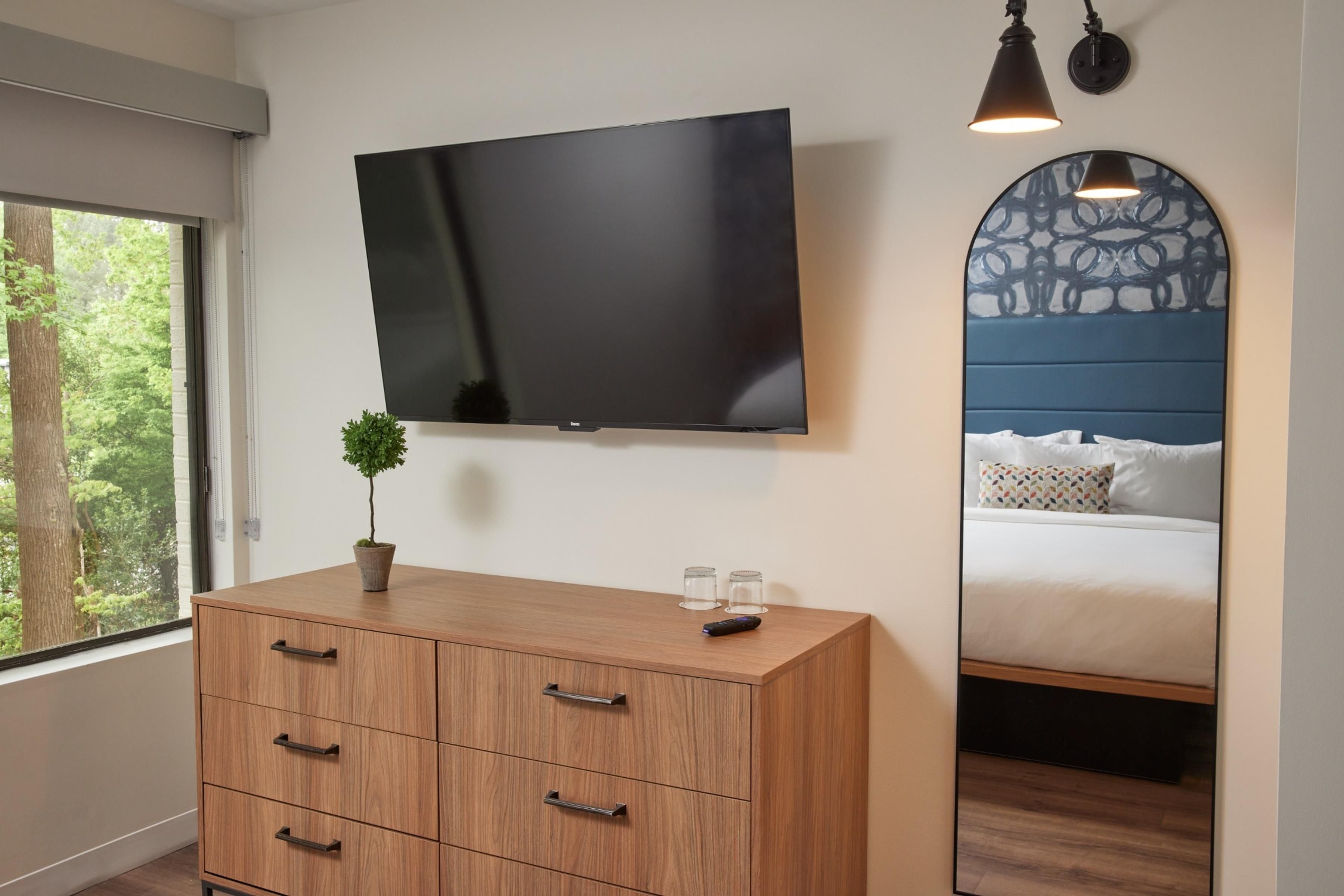 A bedroom with a flat-screen TV mounted on the wall, a wooden dresser with drawers, a small potted plant, and a bed with a patterned headboard.