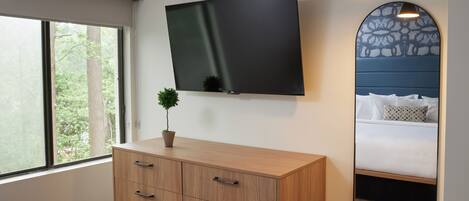 A modern bedroom with a large flat-screen TV mounted on the wall, a wooden dresser with drawers, a bedside mirror, and a window with a view of trees.