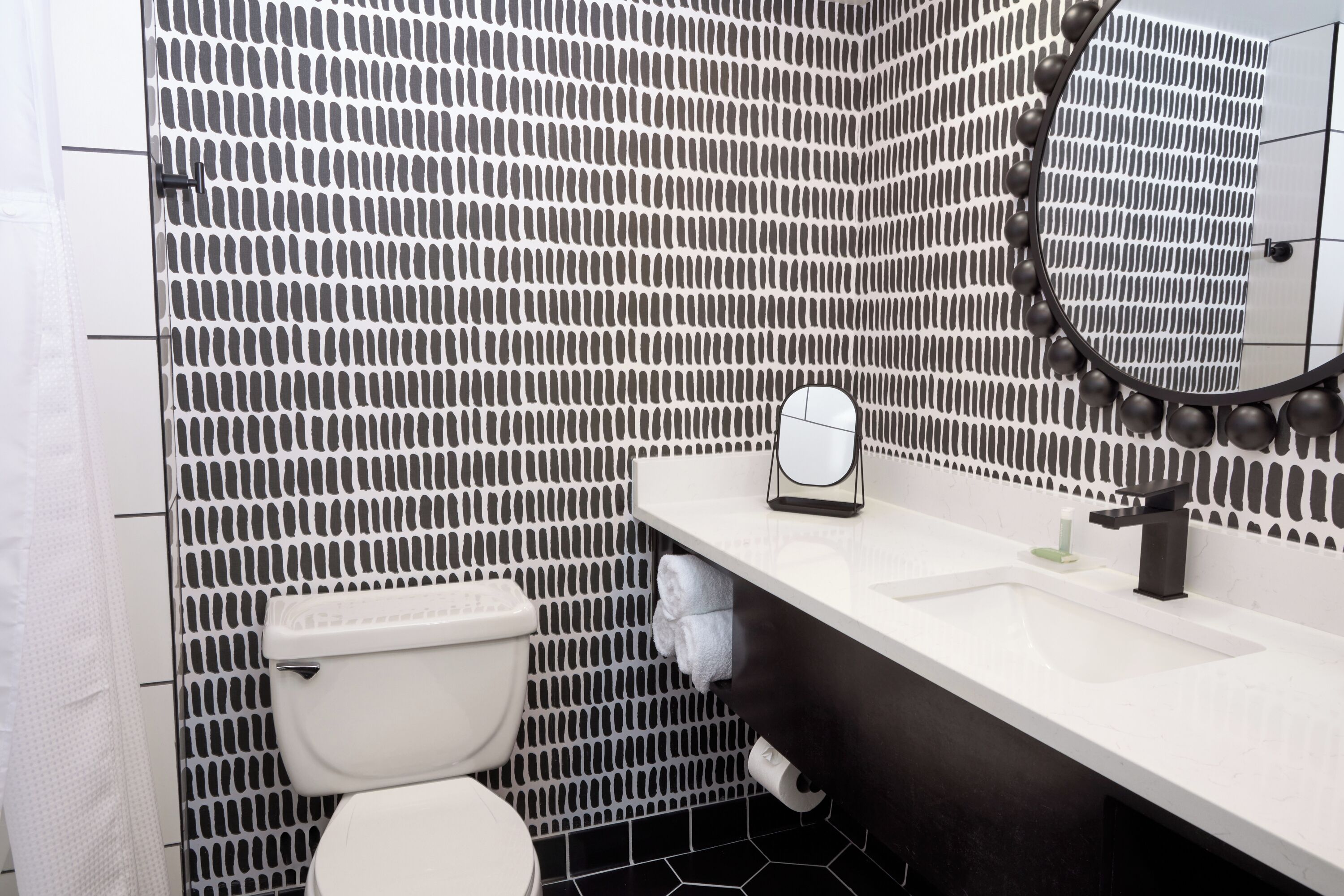 Un baño con pared a cuadros blancos y negros, inodoro blanco, lavamanos con espejo y un perchero para toallas.