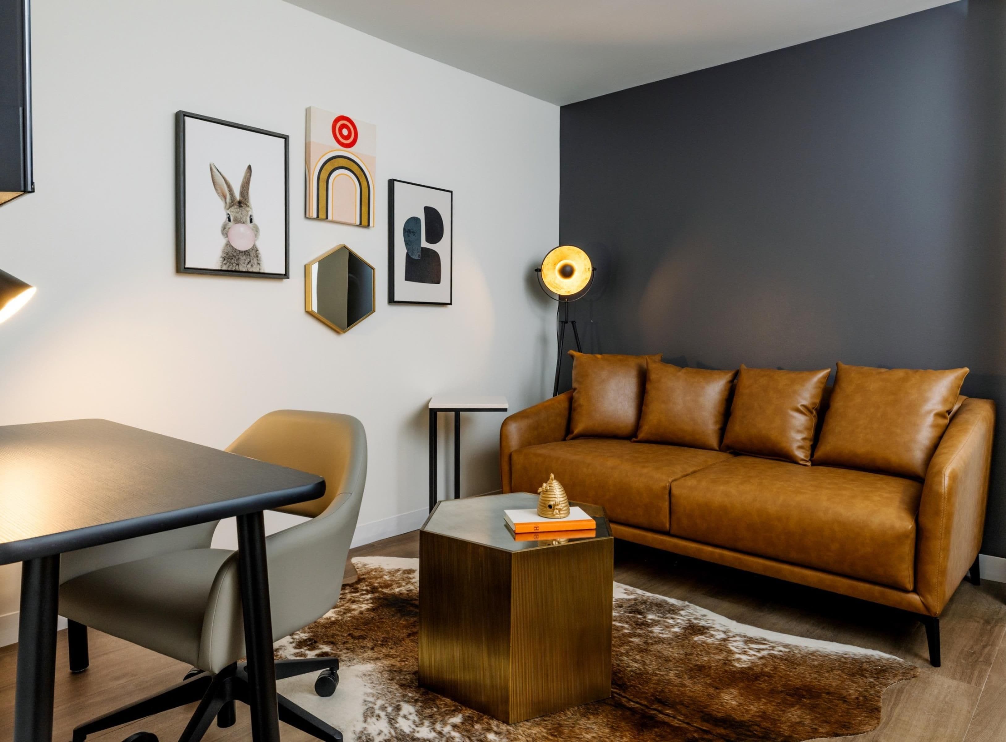 A modern living room with a brown leather sofa, a round table, a desk, and a chair. There are framed pictures on the wall.