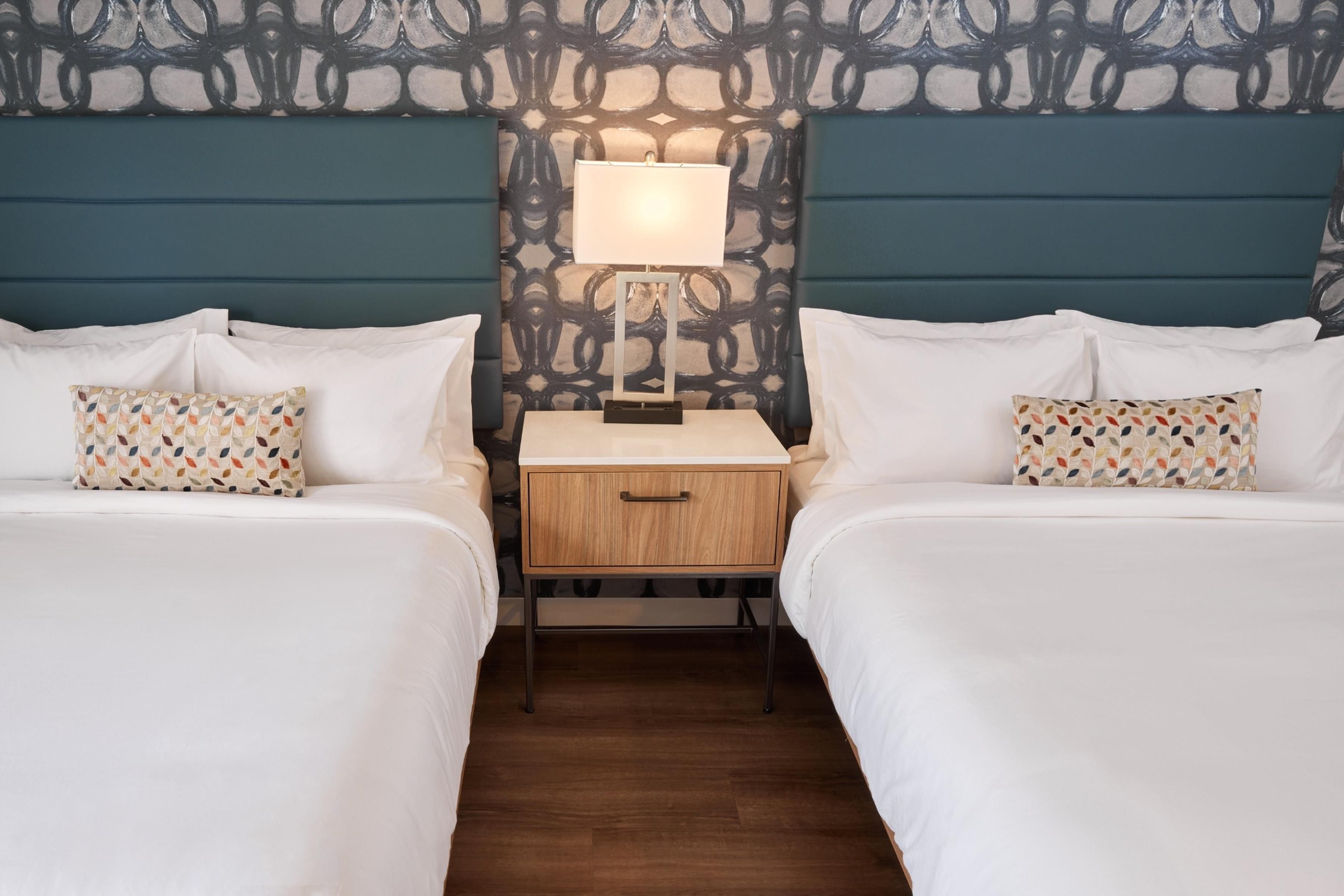 Una habitación de hotel con dos camas, una mesita de noche, una lámpara y un cabecero estampado.