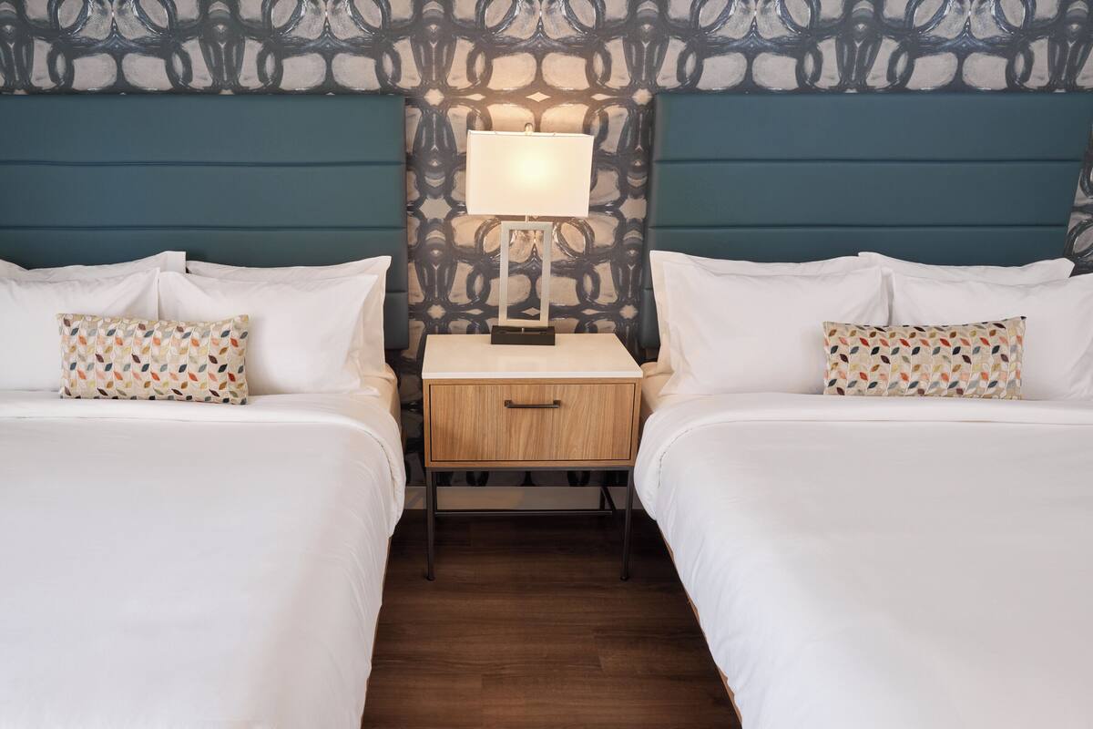 Hotellihuone, jossa on kaksi sänkyä, yöpöytä lampun kanssa ja kuviollinen sängynpääty.