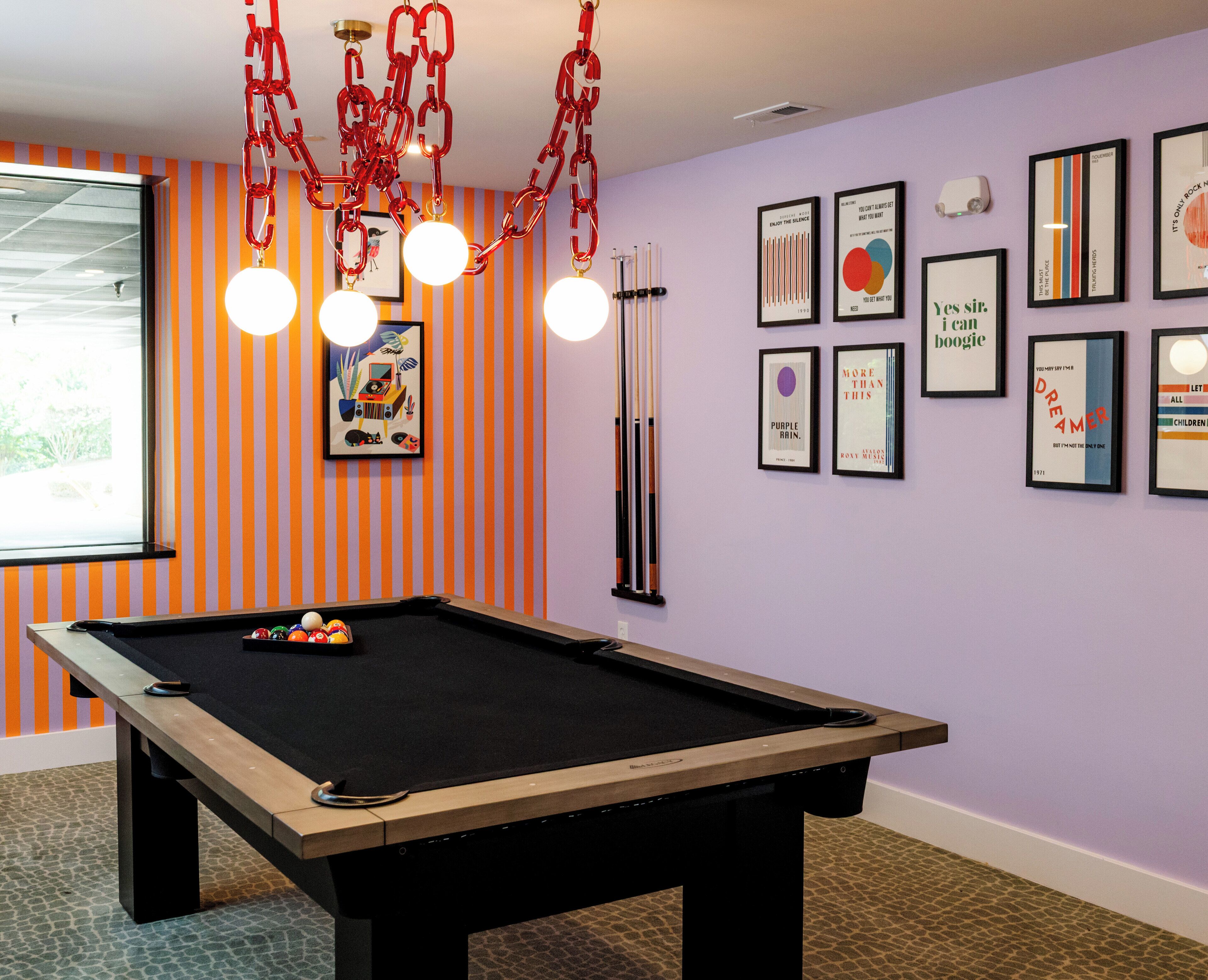 Una mesa de billar con estructura de madera y fieltro negro, rodeada de paredes a rayas naranjas y una pared morada con carteles enmarcados.