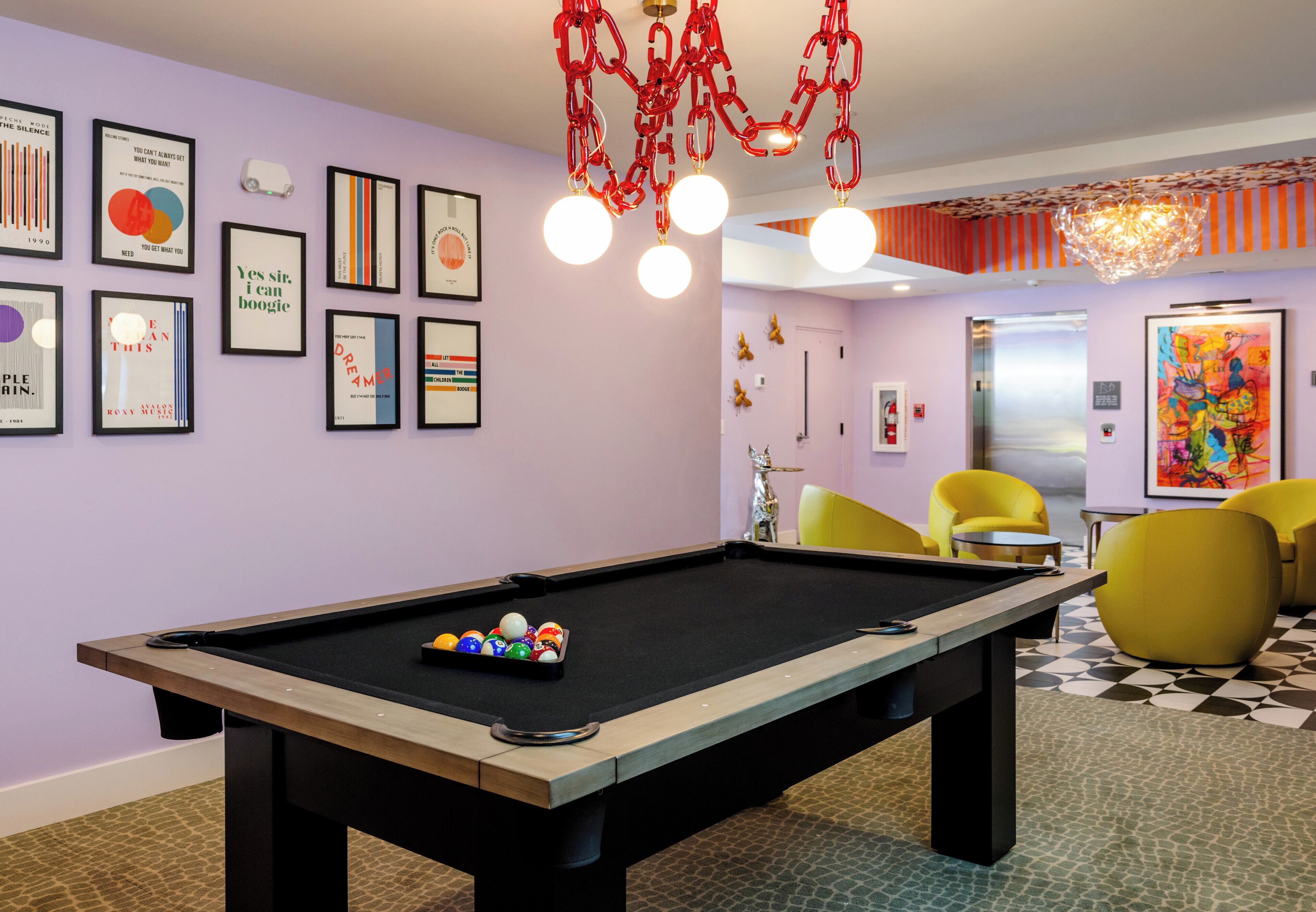 A pool table with balls, a billiards cue, and a pool table cover. There are framed artworks on the wall, a chandelier, and yellow chairs.