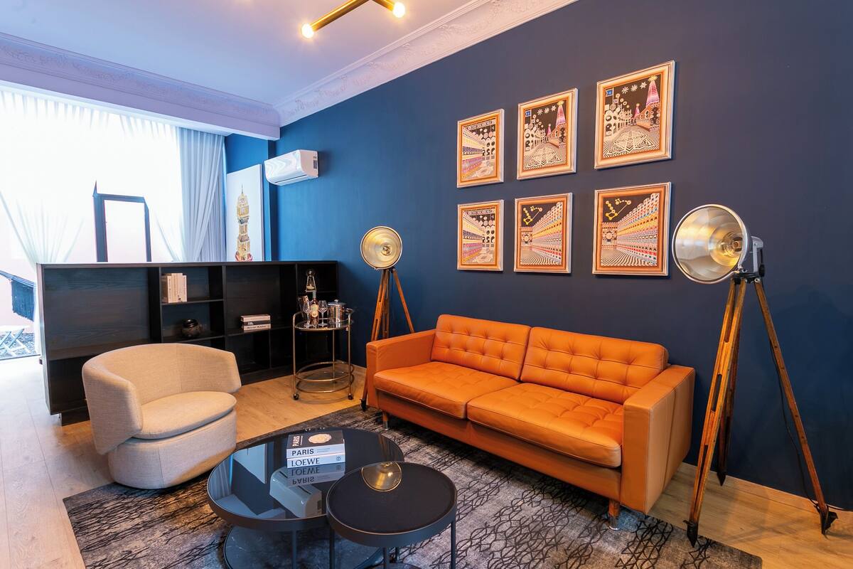 A modern living room with a blue accent wall, a beige armchair, an orange sofa, a glass coffee table, and a bookshelf.