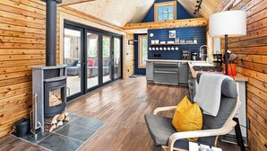 Un acogedor interior de cabaña con estufa de leña, zona de cocina y espacio para sentarse con una silla y cojines.