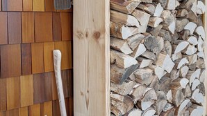 Un montón de leña apoyado contra una pared de madera, un cubo con una pala y una tabla de madera recostada contra la pared.