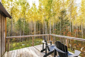 A wooden deck with two chairs and a table, overlooking a forest with autumn foliage.