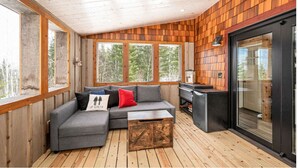 Interior acogedor de una cabaña con una terraza de madera, un sofá seccional gris con cojines rojos y negros, y una estación de parrilla negra.