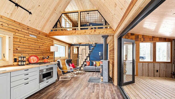 Amplio interior estilo loft con techo alto, paredes de madera, una zona de cocina, un área de estar con sofá y sillas, y una escalera que lleva a un nivel superior.