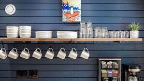 Una zona de cocina con tazas blancas colgando, pilas de platos blancos y una máquina de café.