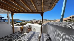 A sunlit terrace with a stone floor, a wooden pergola, and a view of the sea and hills.