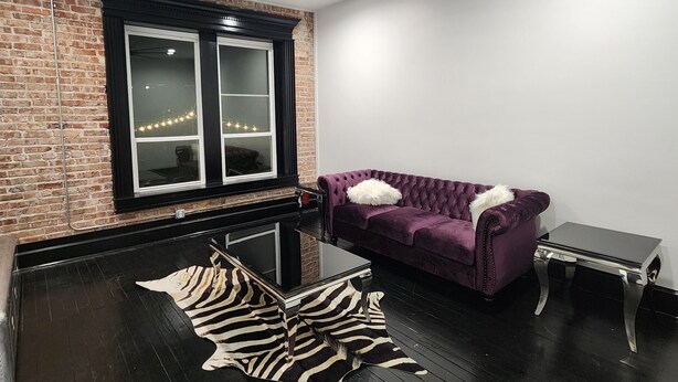 Ein Zimmer mit einer Ziegelwand, einem großen Fenster mit Vorhängen, einem violetten Samtsofa, einem Zebra-Muster Teppich und einem schwarzen Deckenventilator.