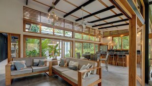 A spacious living area with a wooden sofa set, large windows, and a kitchen area visible through the open space. - Villa Opalo (Cahuita)