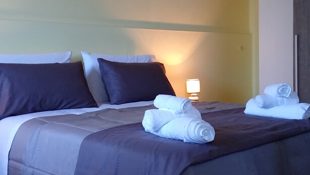 Una cama bien tendida con ropa de cama oscura, una lámpara en la mesita de noche y toallas enrolladas sobre la cama.