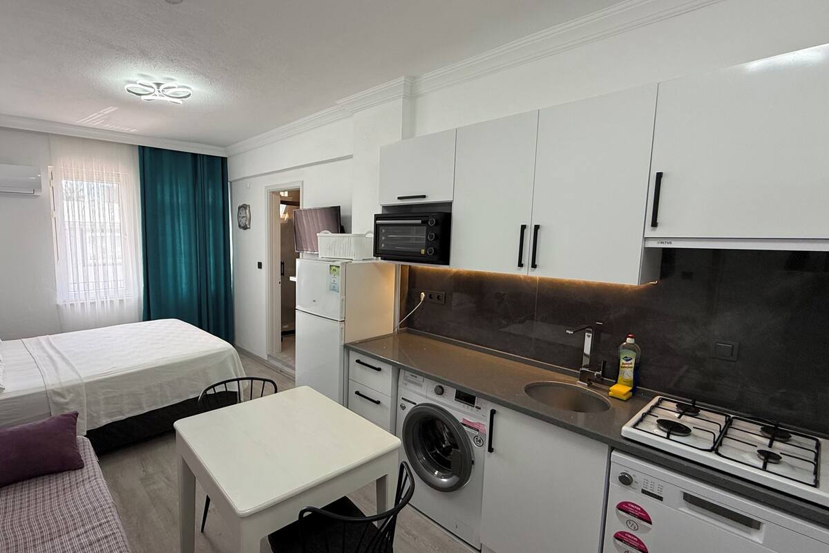 A compact hotel room with a bed, a small table, a washing machine, a stove, and a kitchenette.
