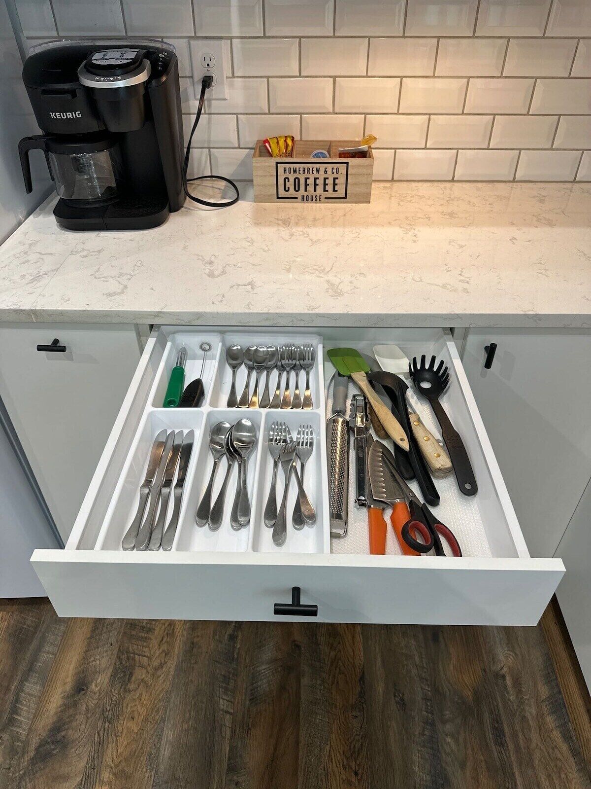 Una cocina con una cafetera Keurig, una caja de café con la etiqueta 'Homebrew & Co. Coffee House', y un cajón que contiene varios utensilios y herramientas de cocina.