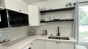 A modern kitchen with white cabinetry, a black microwave, a stainless steel sink, and a window with a view of greenery. - The Loft (Kennesaw)