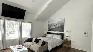 A modern bedroom with a sloped ceiling, a large bed, a sofa, a TV, and a cityscape painting. - The Loft (Kennesaw)