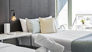 Ein Hotelzimmer mit zwei Betten, einem Nachttisch mit Lampe und Blick auf die Stadt durch ein großes Fenster.