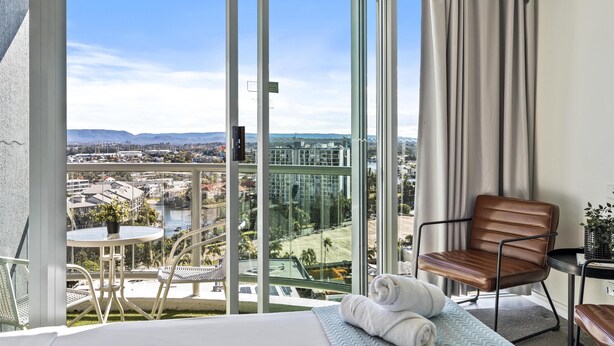 Ein Hotelzimmer mit großem Balkon, einem Bett, einem Sessel, einem kleinen Tisch und Blick auf Stadt und Berge.