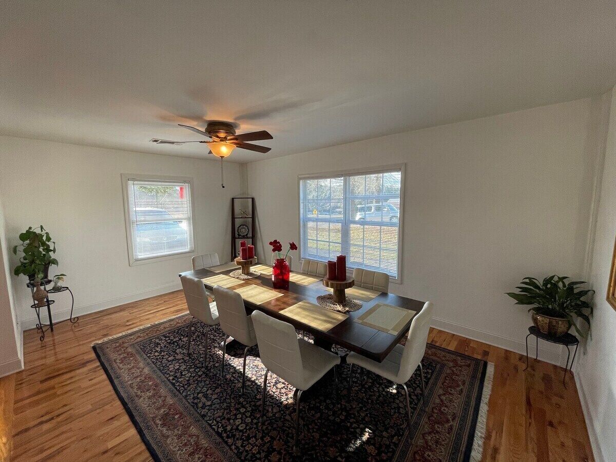 Une salle à manger avec une table en bois, des chaises blanches, un ventilateur de plafond et une grande fenêtre.