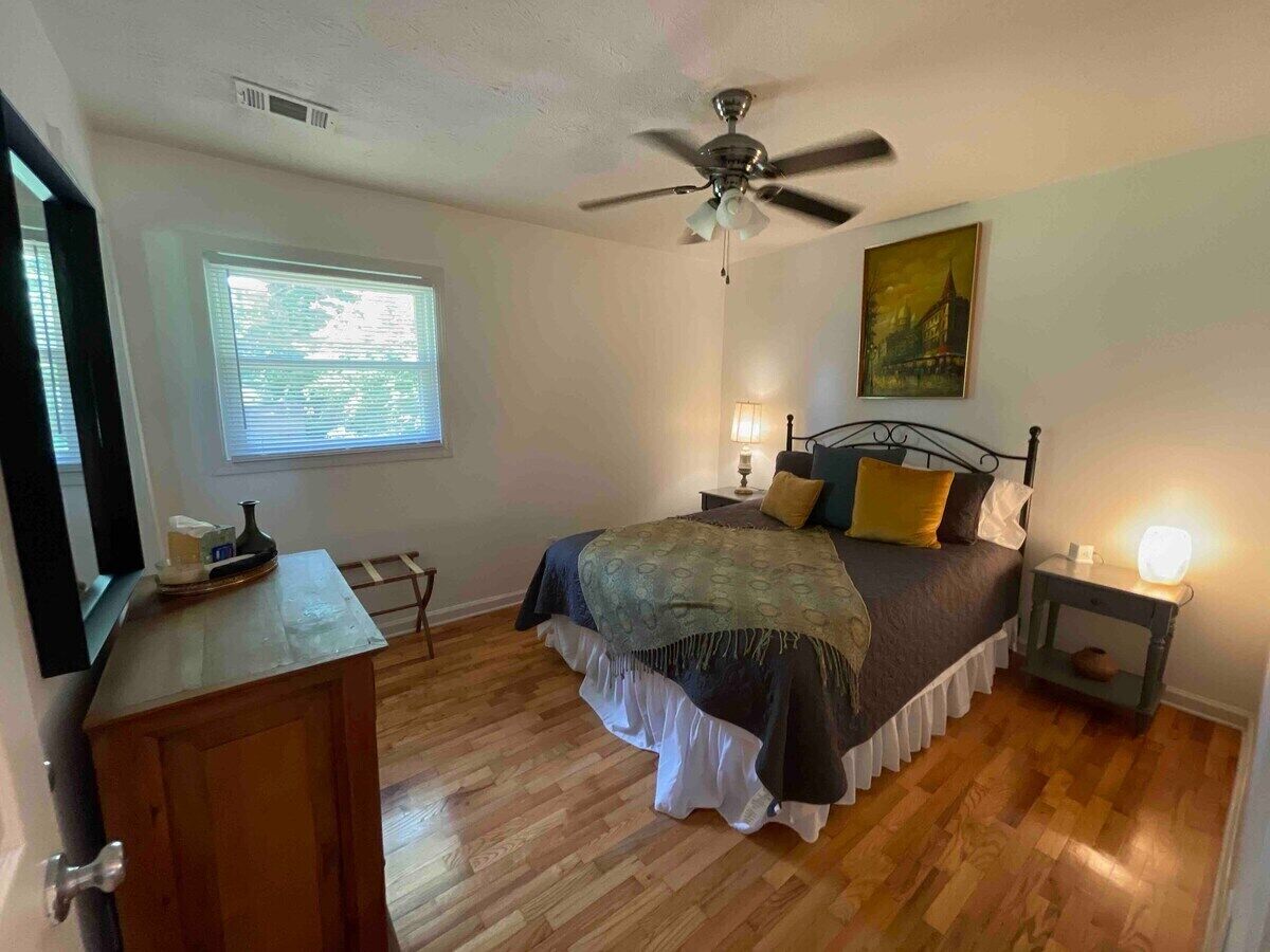 A bedroom with a bed, bedside tables, a ceiling fan, a window with blinds, and a painting on the wall.