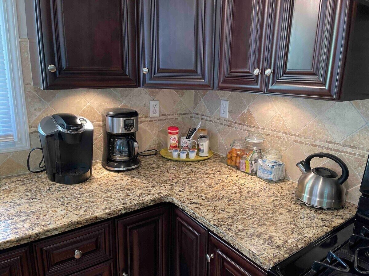 A kitchen with dark wooden cabinets, a granite countertop, a coffee maker, a kettle, and various kitchen utensils and containers.