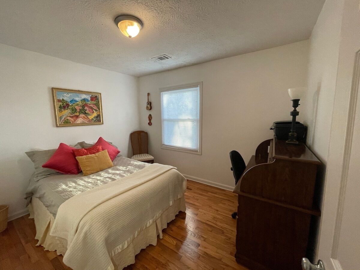Un dormitorio con cama, escritorio, silla, ventana y un cuadro en la pared.