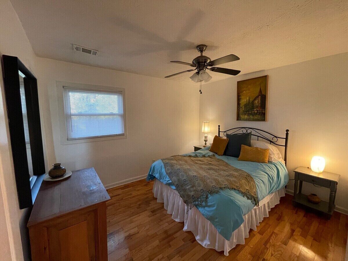A bedroom with a bed, bedside lamps, a ceiling fan, a window with blinds, a wooden dresser, and a painting on the wall.