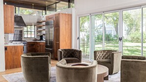 Une cuisine moderne avec des meubles en bois et des appareils en acier inoxydable, un coin repas doté de fauteuils confortables et d’une table ronde, et de grandes portes-fenêtres coulissantes donnant sur un espace extérieur verdoyant.